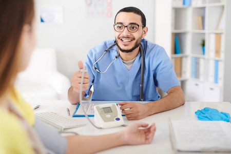 年轻的中东医生戴着眼镜坐在办公室办公桌和测量女性患者血压的肖像