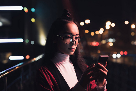 迷人的亚洲女孩在勃艮第和服和眼镜使用智能手机在街上与霓虹灯在晚上, 未来概念的城市