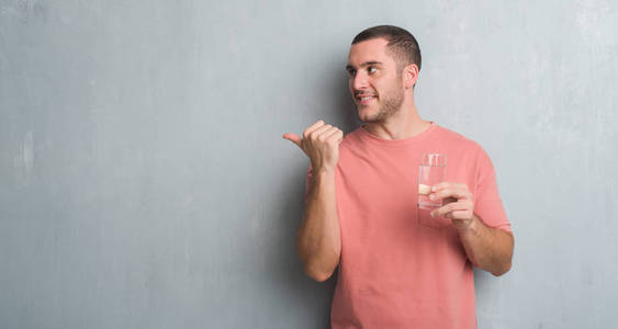 年轻的白种人在灰色垃圾墙壁喝一杯水指向和显示与拇指到边与愉快的面孔微笑