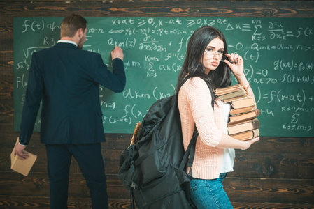 黑头发的学生堆着书和沉重的背包拿着她的眼镜。大学女生站在绿板前教授正在写公式