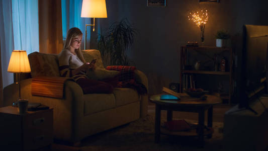 在晚上美丽的年轻女子在沙发上放松在她舒适的客厅里。她用她的智能手机