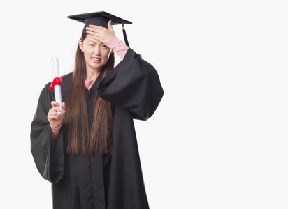 年轻的中国女子身穿毕业制服持纸度强调用手在头上, 感到羞愧和惊讶的脸, 愤怒和沮丧。对错误的恐惧和不安