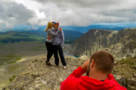 一对爱的情侣站在山上亲吻在摄影师面前的背景下沐浴在阳光, 丘陵, 蓝色湖泊与冰川, 岩石与雪和白云