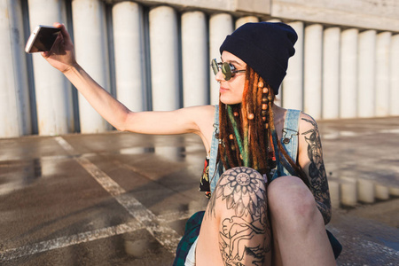 年轻的女孩与纹身和长发绺在蓝色帽子使用智能手机的背景下的混凝土墙