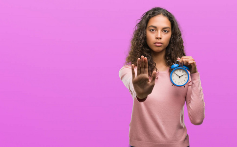 年轻的西班牙妇女手持闹钟与张开手做停止标志认真和自信的表达, 防御姿态