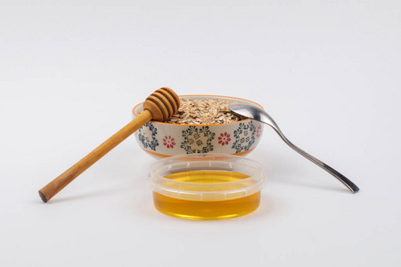 一个充满燕麦片的陶瓷碗和一个带有蜂蜜的容器, 一个不锈钢勺子和一个白色背景的蜂蜜北斗