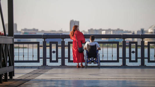 一个漂亮的红头发的女孩穿着红色的连衣裙和一个坐在轮椅上的男人在路堤的边缘, 欣赏的看法
