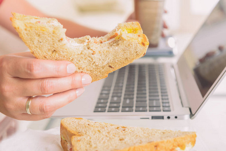 在使用笔记本电脑时吃早餐三明治和喝咖啡的妇女