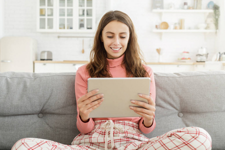 年轻漂亮的微笑的青少年女孩坐在沙发上, 手持数字平板电脑与双手, 看电影或使用社交媒体