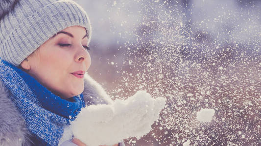 漂亮的年轻女子吹雪玩。带绒球和蓝色围巾的灰色豆帽的女性