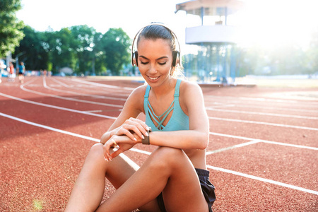 微笑年轻的 sportsgirl 在耳机穿着运动服坐在体育场, 看着她的 smartwatch