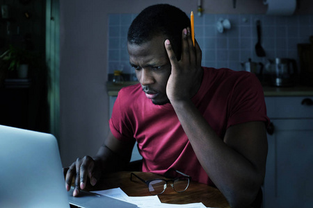 年轻的非洲裔美国男子坐在家里晚上在笔记本电脑与焦虑的脸试图检查重要数据, 看失望, 不满意当前情况