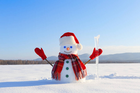 雪人手里戴着格子围巾红帽子手套和冰柱。美丽的风景与山的背景。被雪覆盖的田野。寒冷的冬日