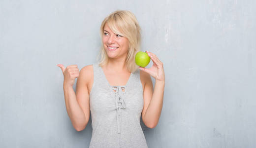 成人白种妇女在垃圾灰色墙壁吃绿色苹果指向和显示与拇指由边与愉快的面孔微笑