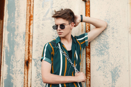 英俊的年轻模特男子与时尚太阳镜的发型和时尚的沙滩衬衫附近的老式金属生锈的墙壁