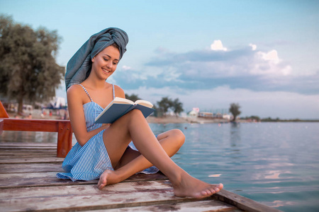 美丽的年轻女子穿着礼服, 头上戴着毛巾, 坐在水边的木墩上, 一边看书一边欣赏