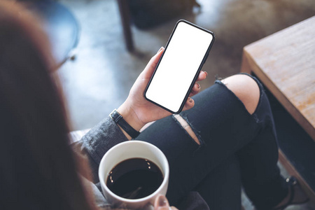 在咖啡馆里喝咖啡时, 一个女人坐在交叉腿上, 手持黑色手机, 大腿上空白的白色桌面屏幕。