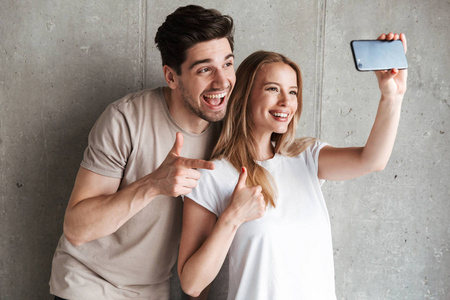 照片的两个微笑的人男人和女人20s 采取自拍照片上的手机, 而手势在相机隔离在混凝土灰色墙室内