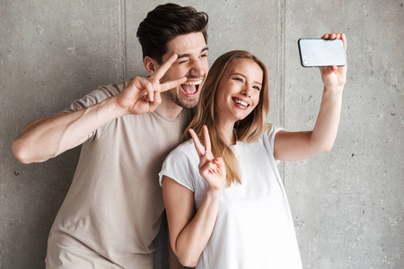 美丽的情侣和女人的形象自拍照片上的智能手机, 而显示和平标志隔离在混凝土灰色墙室内