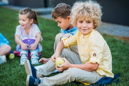可爱的卷发小学生与苹果坐在草地上与同学