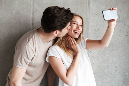 英俊的男人的形象亲吻漂亮的女人脸颊上, 并采取自拍照片的智能手机隔离在混凝土灰色墙室内
