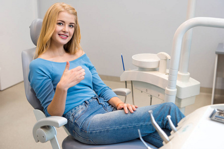 快乐的年轻女子坐在牙医办公室的牙科椅上, 并显示拇指
