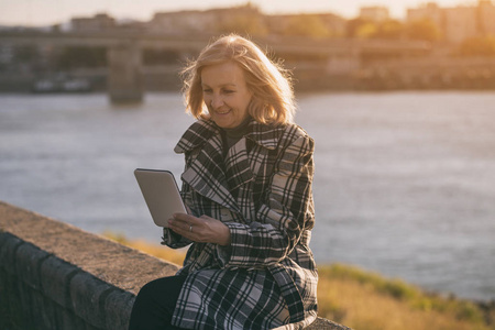 坐在河边时使用数字平板电脑的老年女性。色调的图像