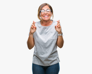 中年资深西班牙裔妇女戴着眼镜在与世隔绝的背景下微笑着交叉手指, 希望和眼睛闭上。运气和迷信概念