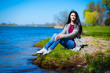 在阳光明媚的日子里, 穿着牛仔裤的漂亮女孩坐在河岸上.照片