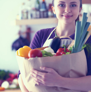 年轻女子捧着与蔬菜站在厨房里的食品杂货店购物袋