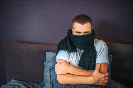 生病的年轻人坐在床上, 用围巾捂住嘴。房间里很冷。这家伙的手是交叉的。他感冒了。年轻人遭受