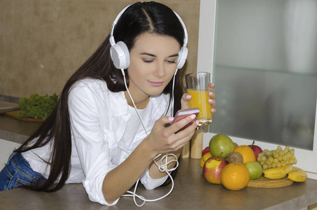 耳机里的女孩听音乐和饮料新鲜榨橙汁, 在厨房里的早晨
