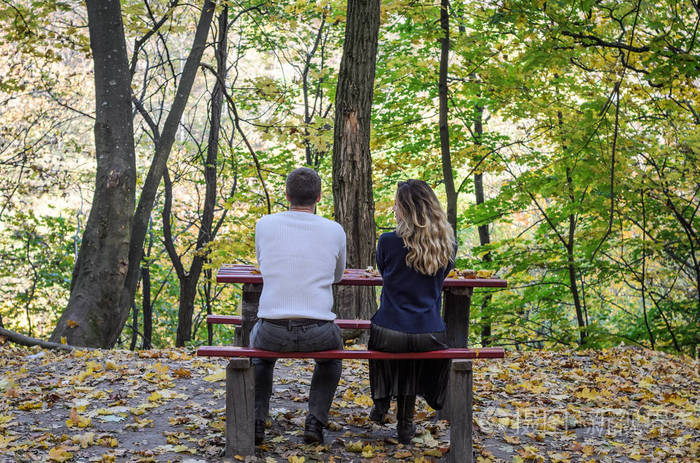 相爱的情侣坐在秋天公园的长凳上的黄落叶之间