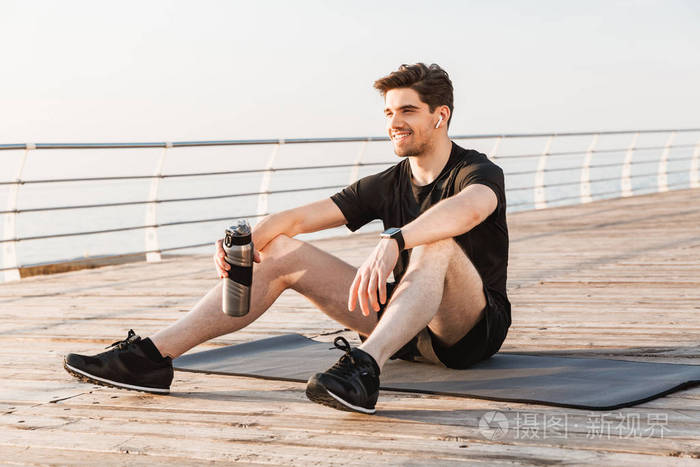 英俊年轻运动员的照片坐在户外与瓶装水在海滩上听音乐与耳机
