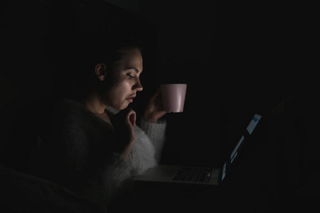 年轻的女孩坐在床上深夜, 在黑暗中, 看着笔记本电脑上的电影