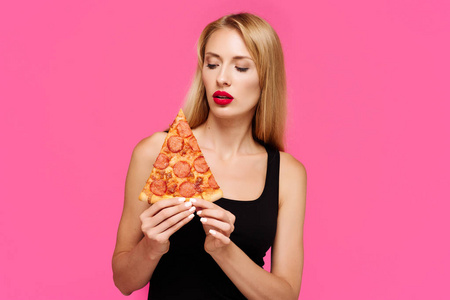 年轻苗条的女孩与粉红色的背景, 在她手中的比萨饼。不健康脂肪垃圾食品的概念