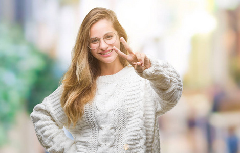 年轻美丽的金发女子穿着冬季毛衣和太阳镜在孤立的背景微笑着看着相机显示手指做胜利标志。第二个