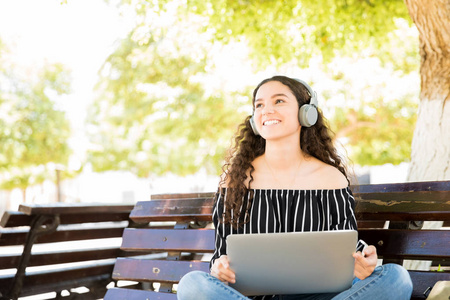 可爱的拉丁少女肖像坐在公园长椅上, 笔记本电脑看着相机和微笑
