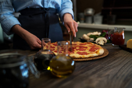 妇女削减新鲜烘焙自制比萨饼在乡村厨房的背景。切成切片美味的比萨饼与蘑菇和火腿。木桌上的奶酪和西红柿。健康食品, 烹饪理念