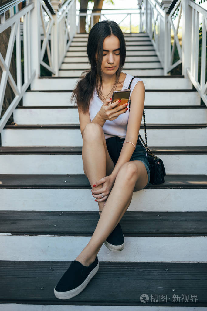 女孩穿着牛仔短裤和白色上衣看着她的移动设备在夏天的一天.