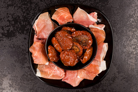 开胃菜来开胃菜。Prosciutoo 火腿和太阳晒干的西红柿在黑盘子上的黑色桌子上。奢华饮食
