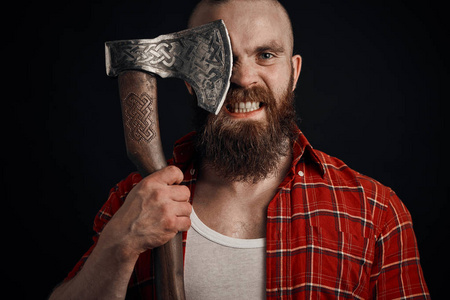 有胡子的男人拿着一把斧头遮住眼睛, 看着黑色背景上的摄像头.照片