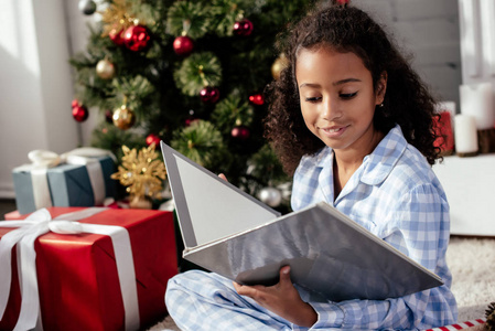 愉快的可爱的非洲裔美国人的孩子在睡衣阅读书附近的圣诞树在家