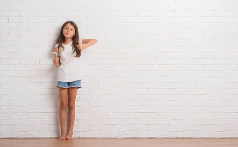 年轻的西班牙裔孩子在白色砖墙吃棒糖糖果在家愉快与大微笑做 ok 标志, 拇指用手指, 优秀标志