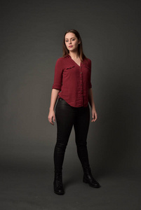 穿红色衬衫和皮裤的黑发女孩的全长肖像。站在姿势, 在灰色的工作室背景