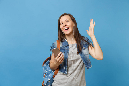 年轻快乐的女学生在牛仔服装与背包, 耳机传播手听音乐举行手机隔离在蓝色背景。大学教育。复制广告空间