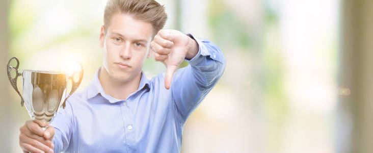 年轻英俊的金发男子拿着一个奖杯与愤怒的脸, 否定的标志显示不喜欢拇指下来, 拒绝概念