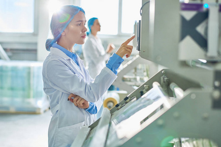 年轻女工穿着实验室大衣和头发盖站立的侧面图清洁生产车间控制面板上的按下按钮