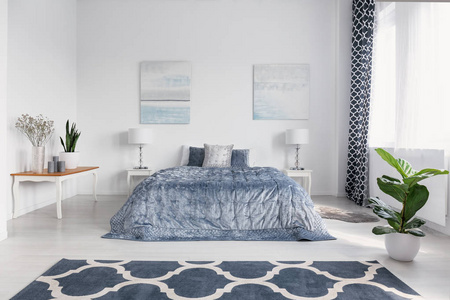 优雅的卧室内饰与大舒适的床, 蓝色床上用品, 墙上的画和地板上的图案地毯, 真实的照片