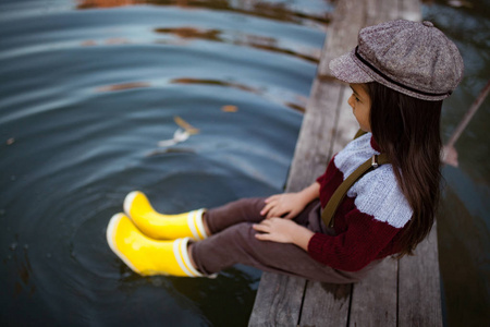 戴黄色橡胶靴和帽子的女孩坐在木桥上, 把腿放在河的背景上的水中